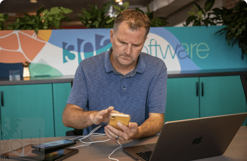 A focused man holding a smartphone connected to a laptop, representing our dedication to mobile application development.
