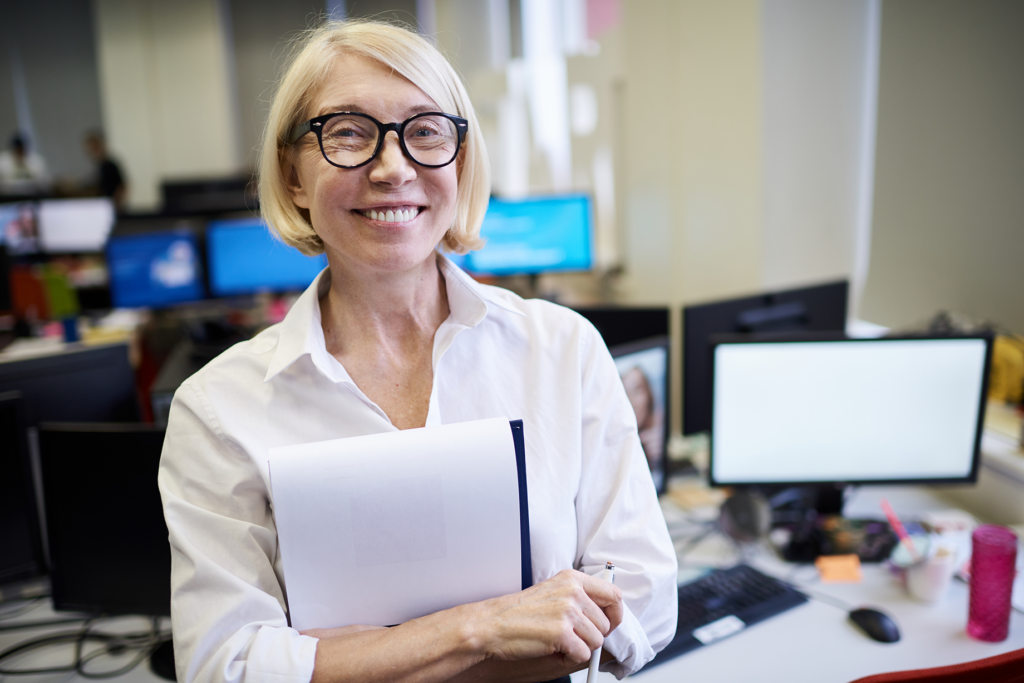 A smiling female college professor with short blonde hair and glasses, holding a folder in a modern office.